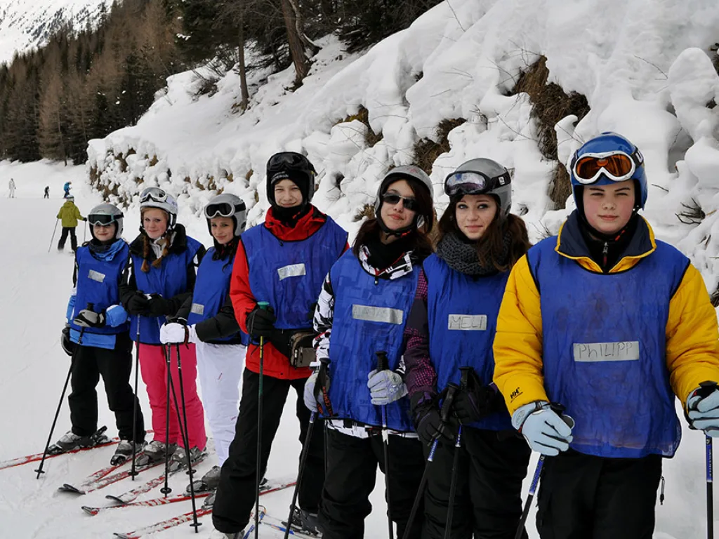 Gruppe in blauen Leibchen steht mit Skiern vor einer Schneewand.